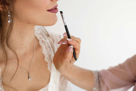 Makeup artist makes makeup to a young bride. A professional makeup artist applies lipstick to a woman's lips with a brush. Makeup artist's hands near the bride's face close upの写真素材