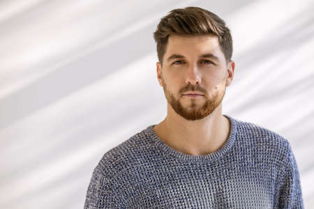 Photo portrait of a handsome man with a fashionable hairstyle and beard on a white background with shadows. A serious man is wearing a gray sweater. Young man staring intently at the cameraの写真素材