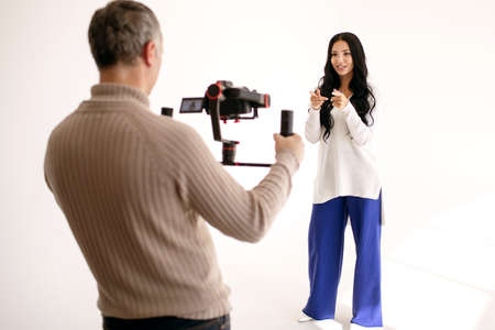 A young woman blogger shoots a video blog in the studio with the help of a videographer with a professional video camera. Modern work as a blogger or journalist. Popular video blogger gesturesの写真素材