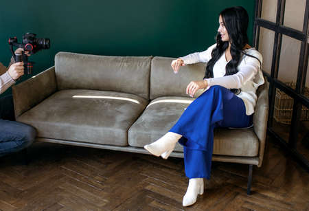 A brunette woman blogger is sitting on a couch indoors and vlogging with a cameraman. Young woman blogger, journalist filming with professional camera and videographerの写真素材