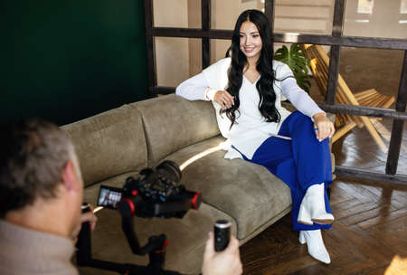 A female blogger sits on a couch indoors and vlogs with a cameraman. Young woman blogger, journalist filming with professional camera and videographerの写真素材