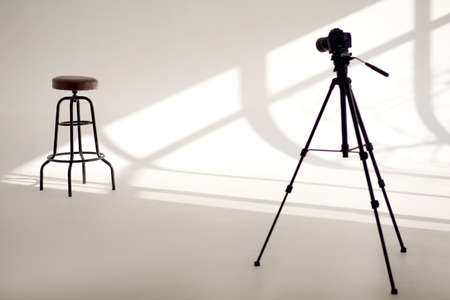 A white photo studio with sun shadows from the windows and one high chair and a video camera on a tripod. Professional photography with bar stool for model and white space with shadowsの写真素材