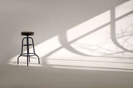 A white room with a shade from the sun through the window and one high bar stool. Minimalism style in horizontal photography. Loneliness concept with one chair and white space with shadowsの写真素材