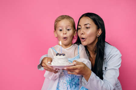 Young beautiful brunette mother and her little daughter are celebrating a birthday. Happy family blowing out candles on daughter's birthday on pink backgroundの写真素材