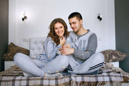 Happy and enamored guy and girl are sitting on the bed and holding a garland with yellow lights. Christmas atmosphere or family celebrations. Young people in love enjoy each otherの写真素材