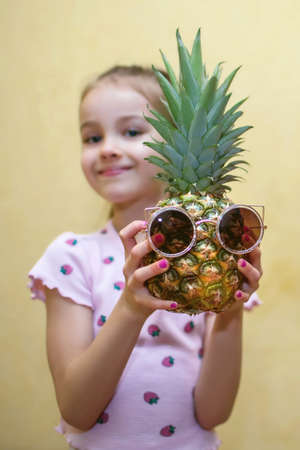 Cute baby girl holding a ripe pineapple in her hands on a yellow background. Pineapple with sunglasses in the form of a human face. Child having fun in a vertical photoの写真素材