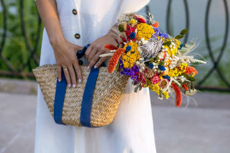 A girl in a white dress holds wild flowers in a wicker straw bag. Vintage eco style concept. Stylish wicker straw bag with dried wildflowers close-upの写真素材