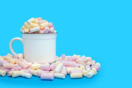 A mug with cocoa or hot chocolate and a lot of colorful marshmallows scattered on a blue background. Sweet airy dessert or addition to hot drinks. Marshmallows with cocoa and free space for textの写真素材