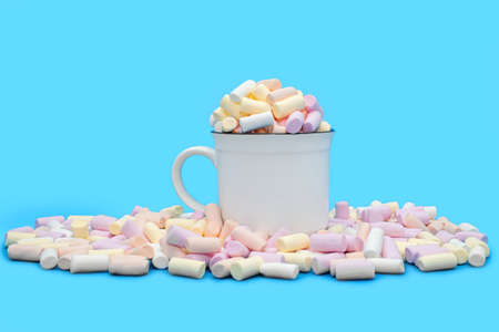 A white mug with cocoa or hot chocolate and many multi-colored marshmallows scattered on a blue background. Sweet airy dessert or addition to hot drinksの写真素材