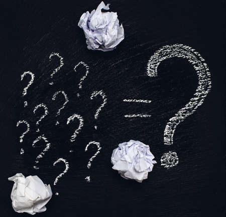 Many small question marks and one large question mark are drawn in white chalk on a rough black...