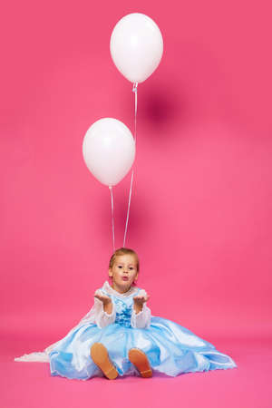 A baby girl in a blue dress sits on a pink background decorated with white balloons. A cute baby smiles and blows a kiss. Vertical photo of a happy child on his birthday. Children's holiday conceptの写真素材