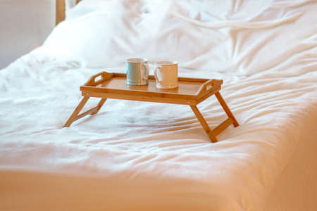 Two mugs with hot drinks on a wooden tray stand on a white bed. Cozy atmosphere or relaxation concept. Breakfast in bed in a cozy roomの写真素材