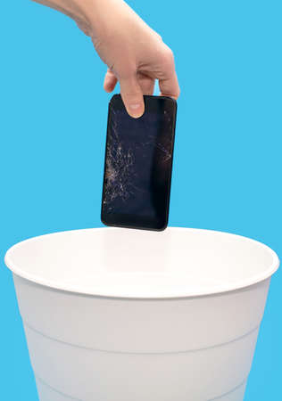 Human throws a broken mobile phone into a trash can on a blue background. Human hand holds a phone with a broken screen over a white trash can and prepares to throw away the broken equipmentの写真素材