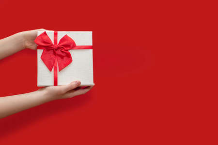 White gift box with a red ribbon in the hands of a man on a red background. Beautiful gift box decorated with ribbon and bow. The concept of giving gifts for birthday or traditional holidaysの写真素材