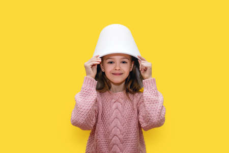 Girl child with a white bucket on her head on a yellow background. Cute baby with a bucket on his...