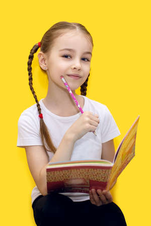 A cute little girl is learning lessons while holding a notebook and a pencil in her hand, or drawing while sitting on a yellow background. A child with pigtails thought while doing a school assignmentの写真素材