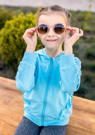 A cute baby girl in a blue jacket and sunglasses sits on a bench in the park and smiles during sunset. Fashionable child with pigtails poses cutely for the photographerの写真素材