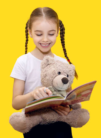 Girl child reads a book and holds a teddy bear on a yellow background. A cute girl with pigtails is learning to read and playing with her teddy bear. children learning concept on vertical photoの写真素材