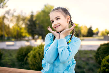 Cute baby girl in a blue jacket with pigtails posing sitting on a bench at sunset. Happy child smiles and has fun in the evening in the park. happy childhood conceptの写真素材