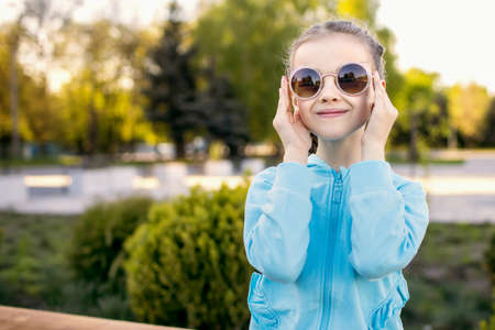 A cute little girl in a blue jacket and sunglasses sits on a park bench and smiles during sunset. Fashionable child with pigtails poses cutely for the photographer. Free space for textの写真素材