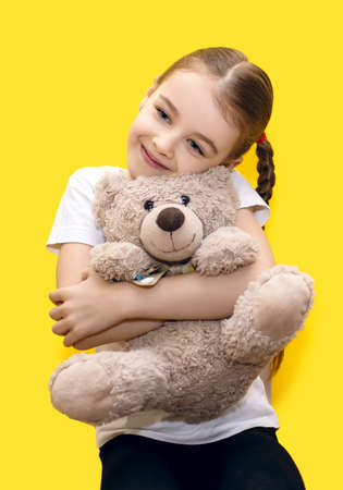 Cute little girl hugging a teddy bear while sitting on a yellow background. A beautiful preschool girl with pigtails smiles and looks good-natured. Happy girl with her favorite toyの写真素材
