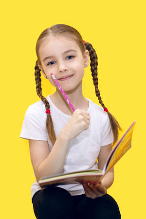A cute little girl is learning lessons while holding a notebook and a pencil in her hand, or drawing while sitting on a yellow background. Child with pigtails is thoughtful and smilingの写真素材