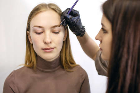 A young woman uses the services of an eyebrow master in a beauty salon. The eyebrow master draws a white outline around the eyebrows of a redhead girl with freckles. beauty and appearance conceptの写真素材