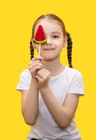 A happy girl holds a candy in the form of a watermelon on a stick and covers part of her face with it on a yellow background. Happy child is having fun. The concept of children's love for sweetsの写真素材
