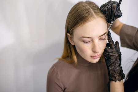 Eyebrow master forms and paints over the eyebrows of a red-haired girl with freckles with a brush. Portrait of a young woman in the process of dyeing her eyebrows. Free space for textの写真素材
