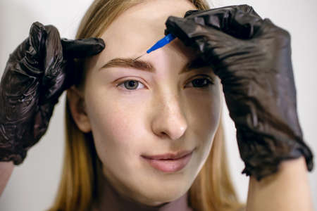 Eyebrow master using a micro brush shapes combining the eyebrows of a red-haired girl with freckles in a beauty salon. Close-up portrait of young woman and hands of stylist in glovesの写真素材