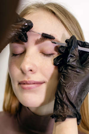 Eyebrow master using a set of tools combs and removes unwanted hairs with tweezers close-up on the client's skin. Red-haired girl with freckles shapes and paints her eyebrows on a vertical photoの写真素材