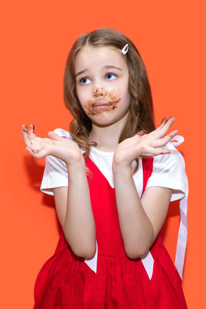 A little girl in a red dress smeared chocolate on her hands and mouth and became upset. Portrait of a child smeared with chocolate on an orange background. The concept of children's love for sweetsの写真素材