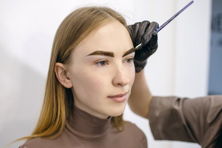 Eyebrow master shapes and paints with a brush the eyebrows of a red-haired girl with freckles in a...