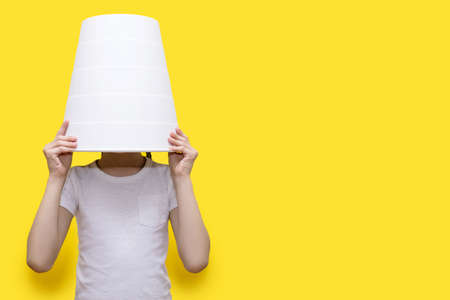 The child put a bucket on his head, sitting on a yellow vertical background. Funny child hidden in a white bucket. The child hides from the parents. Free space for textの写真素材