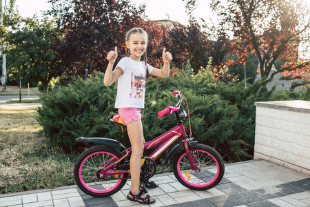 Girl child is dressed in summer clothes and rides a bicycle. Happy smiling child sits on a bike and shows a thumbs up gesture. The girl with pigtails loves to ride a bike. Smiling child on a bicycleの写真素材