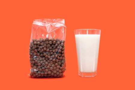 Round chocolate flakes in a transparent package and a glass of milk on an orange background. Packed chocolate flakes in the form of balls and a glass of milk in the center of the imageの写真素材