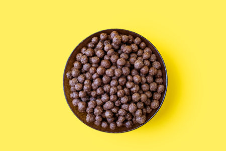 Chocolate corn flakes in a bowl on a yellow isolated background in the center of the image. Flakes with chocolate flavor, close-up top view. American-style energy carbohydrate breakfastの写真素材