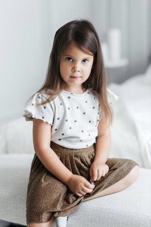 A cute little girl is sitting on a bed in a bright room. A serious child is dressed in a skirt and a T-shirt. Beautiful portrait of a little girl in her bedroomの写真素材