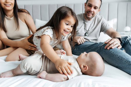 Young parents and their children play in bed in a bright bedroom. Dad, mom, daughter and newborn baby have fun in bed. The eldest daughter plays with her younger newborn brother. happy big familyの写真素材