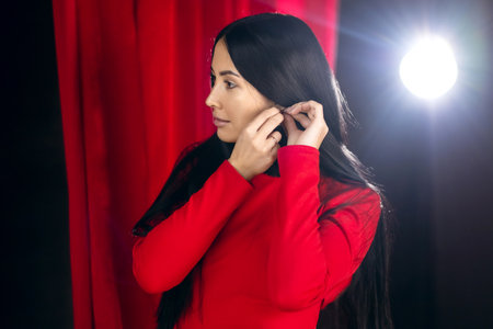 A beautiful brunette woman in a red dress puts on an earring against the background of backlight. Portrait of a cute woman in redの写真素材