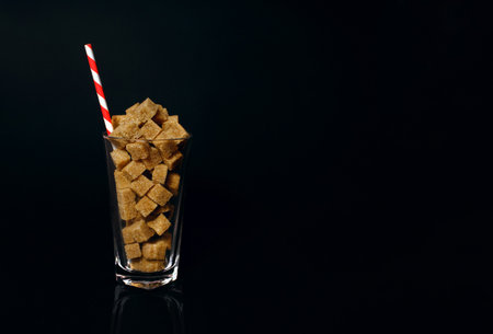 Cane sugar slices in a glass with a red and white drinking straw on a black background. The notion of how much sugar is in sodas, soft drinks, and sweets is dangerous. Free space for text and adsの写真素材