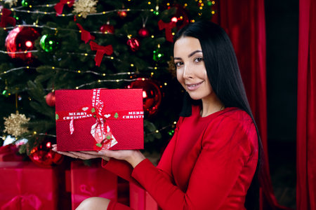 Portrait of a brunette woman with a Christmas gift in a red box on the background of the Christmas tree. Happy brunette rejoices with a Christmas present. gift giving conceptの写真素材