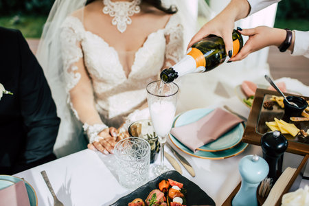 The waiter pours champagne into a glass for the bride at the festive table. Wedding party and drinking alcohol. The waiter serves the table of the newlyweds at the wedding banquetの写真素材