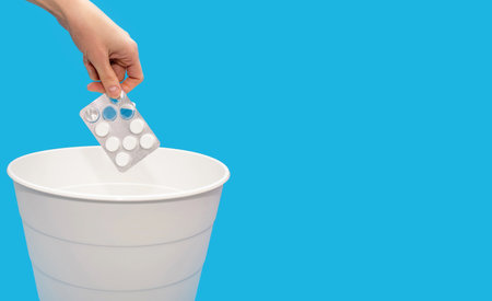 A hand throws pills into a trash can on a blue background with free space m for text. The concept of drug withdrawal or treatment termination. Disposal of expired or unwanted medicinesの写真素材