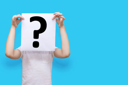 The child holds a piece of paper with a question mark on a blue background and covers his face. The concept of curiosity or indecision. Question mark on white paper with free space for textの写真素材