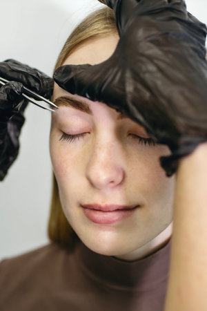 A professional brow stylist uses tweezers to remove excess hair and shape the eyebrows. Fashionable girl takes care of her appearance and looks after her beautyの写真素材