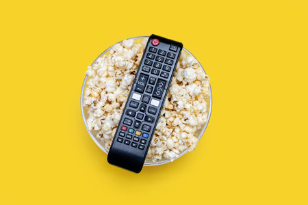 Popcorn in a glass bowl and a TV remote control on a yellow background. A set for relaxing and watching movies or series. Delicious snack of crispy popcorn in a bowlの写真素材