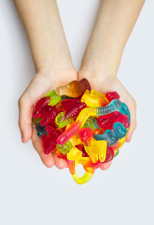 A large pile of multi-colored jelly marmalade candies in the hands of a child on a white background. Variety of tasty bright chewing gums. Various delicious gummy candies in hands close-up top viewの写真素材