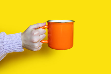 A hand holds an orange cup with a hot drink on a yellow background. The hand of a woman dressed in a sweater holds a ceramic large cup on a bright yellow background. drinking a hot drinkの写真素材
