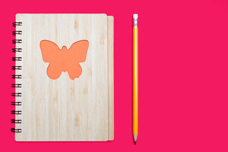 Closed notebook in a bamboo cover with an orange butterfly on a pink background and a pencil. Stationery in the form of a notebook and a pencil on a pink background. Free space for textの写真素材